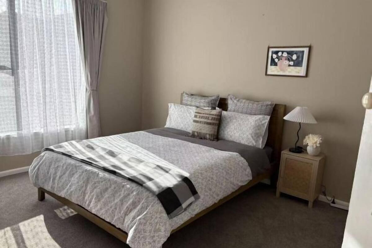 A bedroom with a bed, a bedside table, a lamp, a picture on the wall, and a window with curtains.
