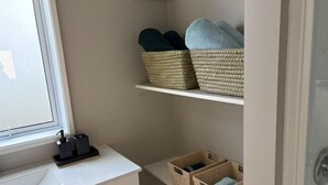 A bathroom with a white countertop, a window, and shelves holding woven baskets with towels. - The Nest on Till (Oamaru)