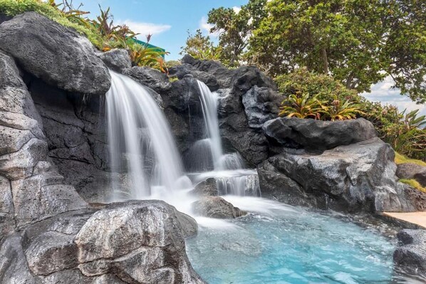 A waterfall cascading over large rocks into a clear pool, surrounded by lush greenery and trees.