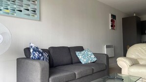 A living room with a grey sofa, two patterned cushions, a glass coffee table, a beige armchair, a wall-mounted fan, and a white air conditioning unit. - Apartment - Sleeps 4, Pets Allowed & Parking (Salford)