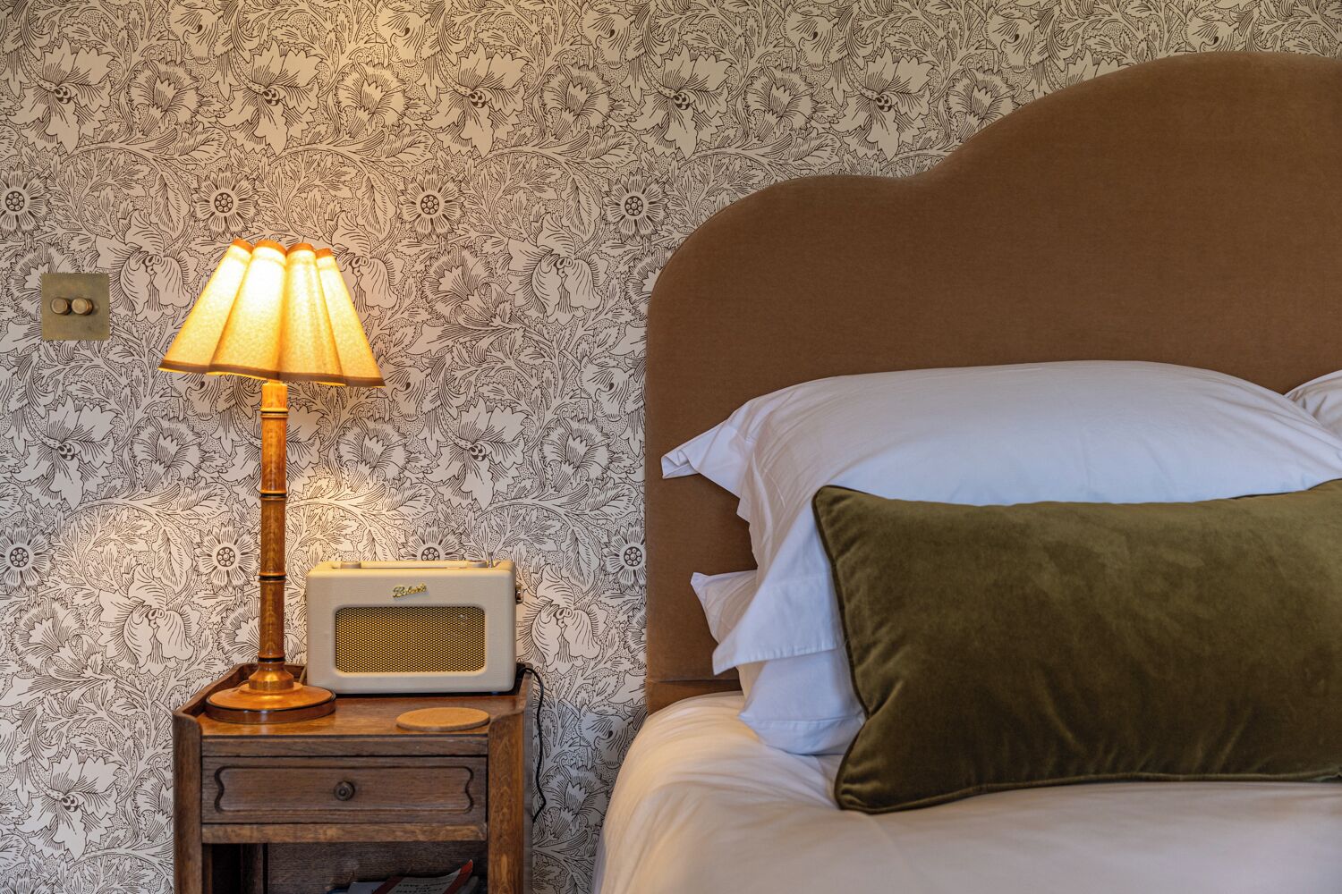 A neatly made bed with white and green pillows, a wooden nightstand with a lamp and radio, and a floral wallpaper background.