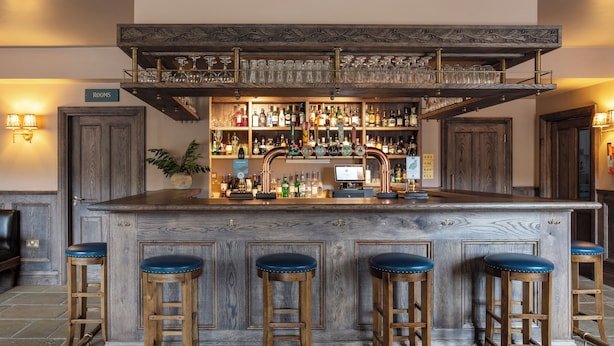 Eine Bar mit einer Holztresen, vier Barhocker, einer Auswahl an Flaschen und einem Schild, das die Zimmer zeigt.