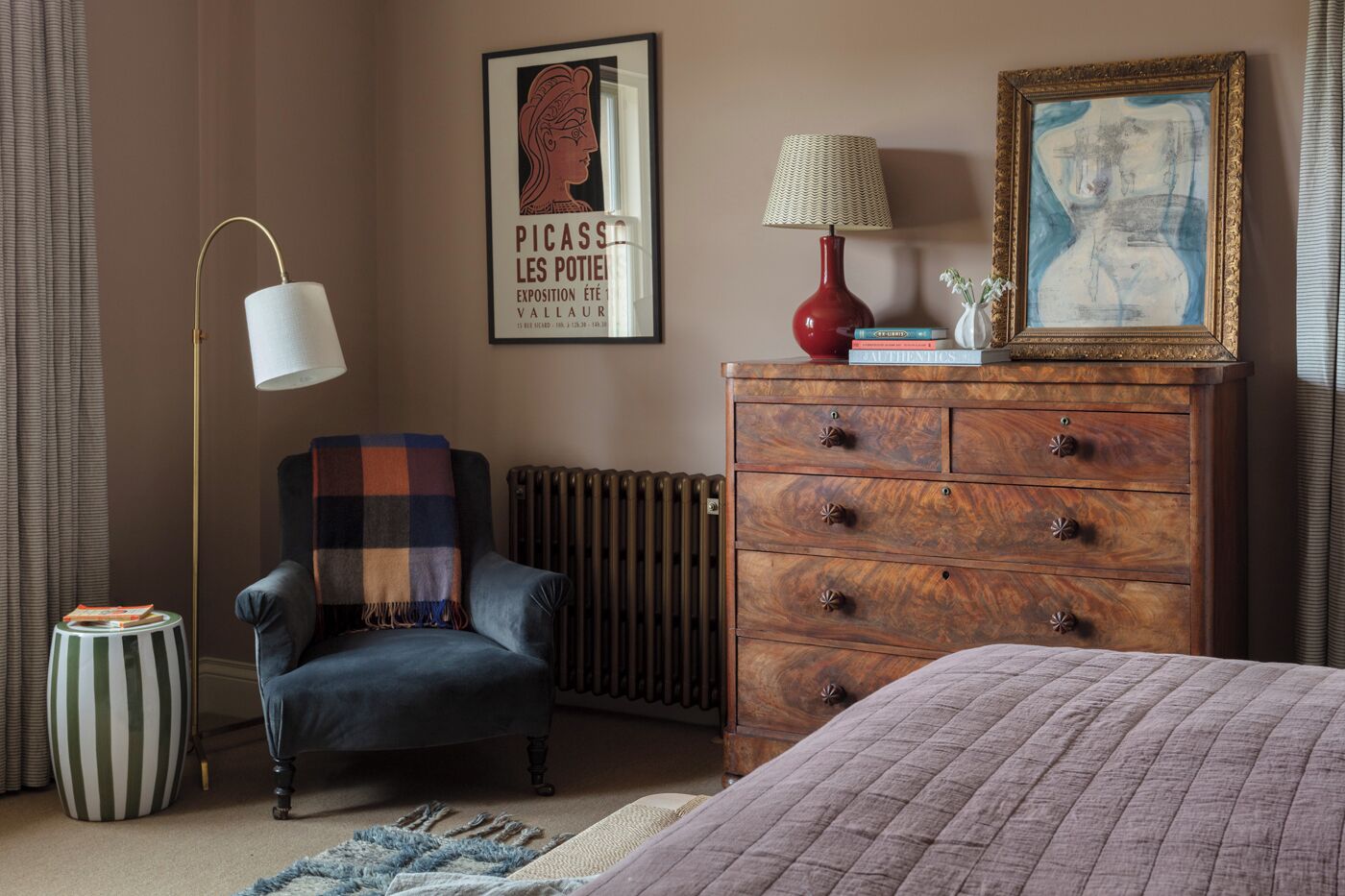 Een slaapkamer met een blauwe fauteuil, een houten dressoir, een radiator en een kunstwerk in een lijst aan de muur.