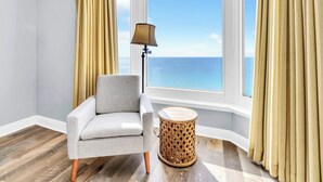 A room with a view of the ocean, a chair, a lamp, and a round wooden side table.