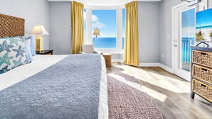 A bedroom with a bed, a window with curtains, a ceiling fan, and a view of the beach.