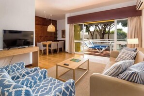 A modern living room with a TV, sofa, coffee table, and a balcony with a pool. - Apartamentos Turisticos Pinhal Da Marina (Vilamoura)