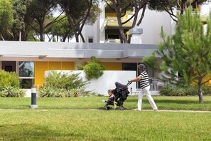 Eine Person schiebt einen Kinderwagen über eine Wiese, im Hintergrund ein modernes Gebäude.