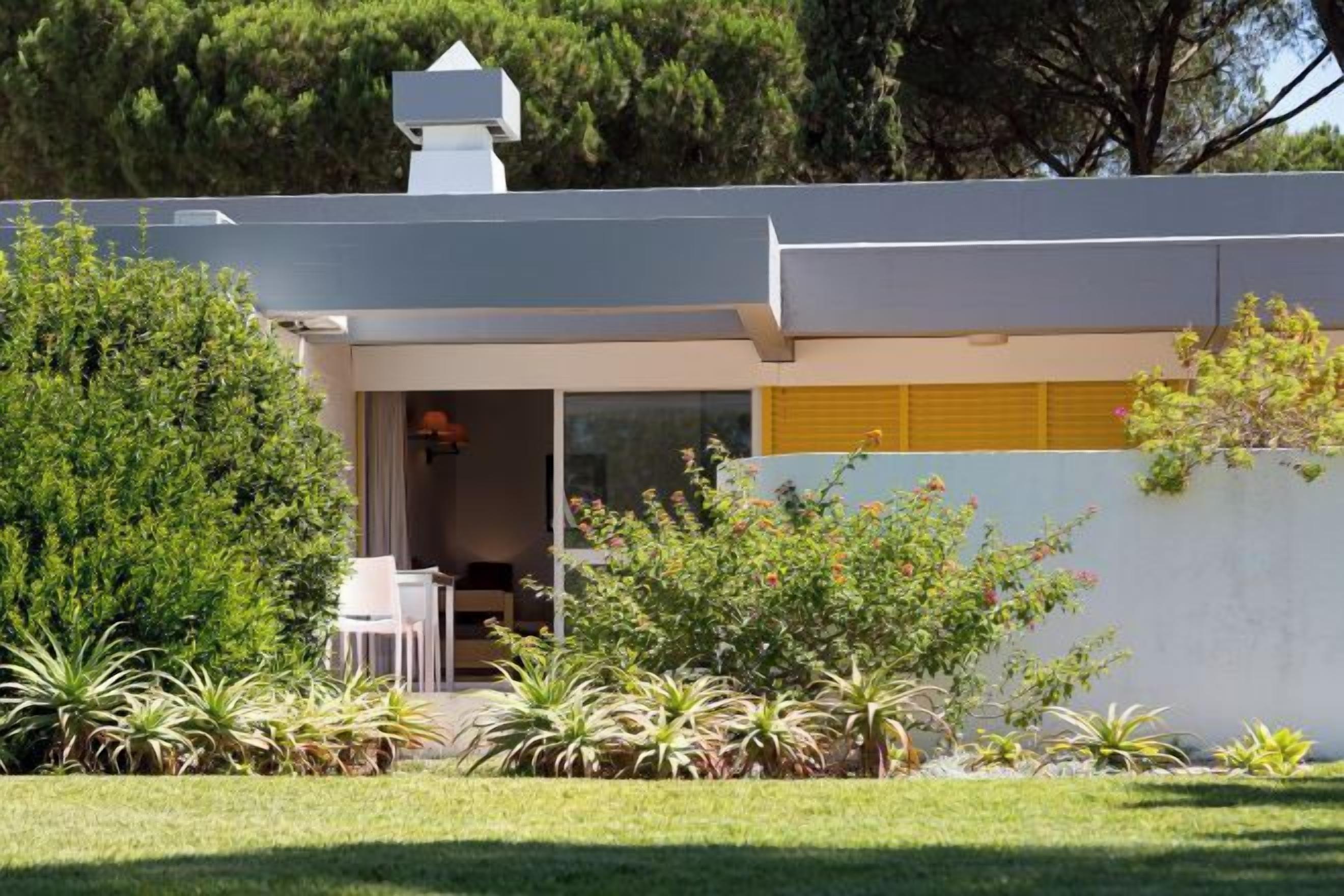 Ein modernes Haus mit überdachter Sitzgelegenheit im Freien, einem sichtbaren Fenster und einem Garten mit verschiedenen Pflanzen.