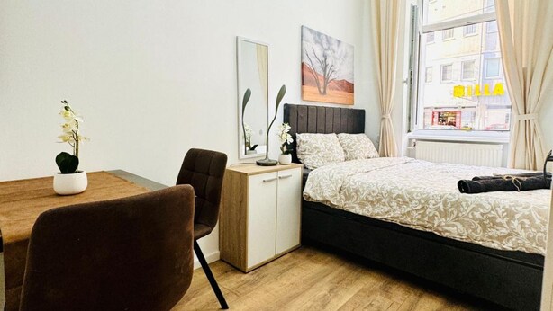 A bedroom with a bed, a desk, a chair, a potted plant, and a window with curtains.