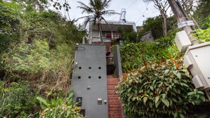 A residential building with a concrete facade, a metal staircase, and a gated entrance surrounded by dense vegetation.
