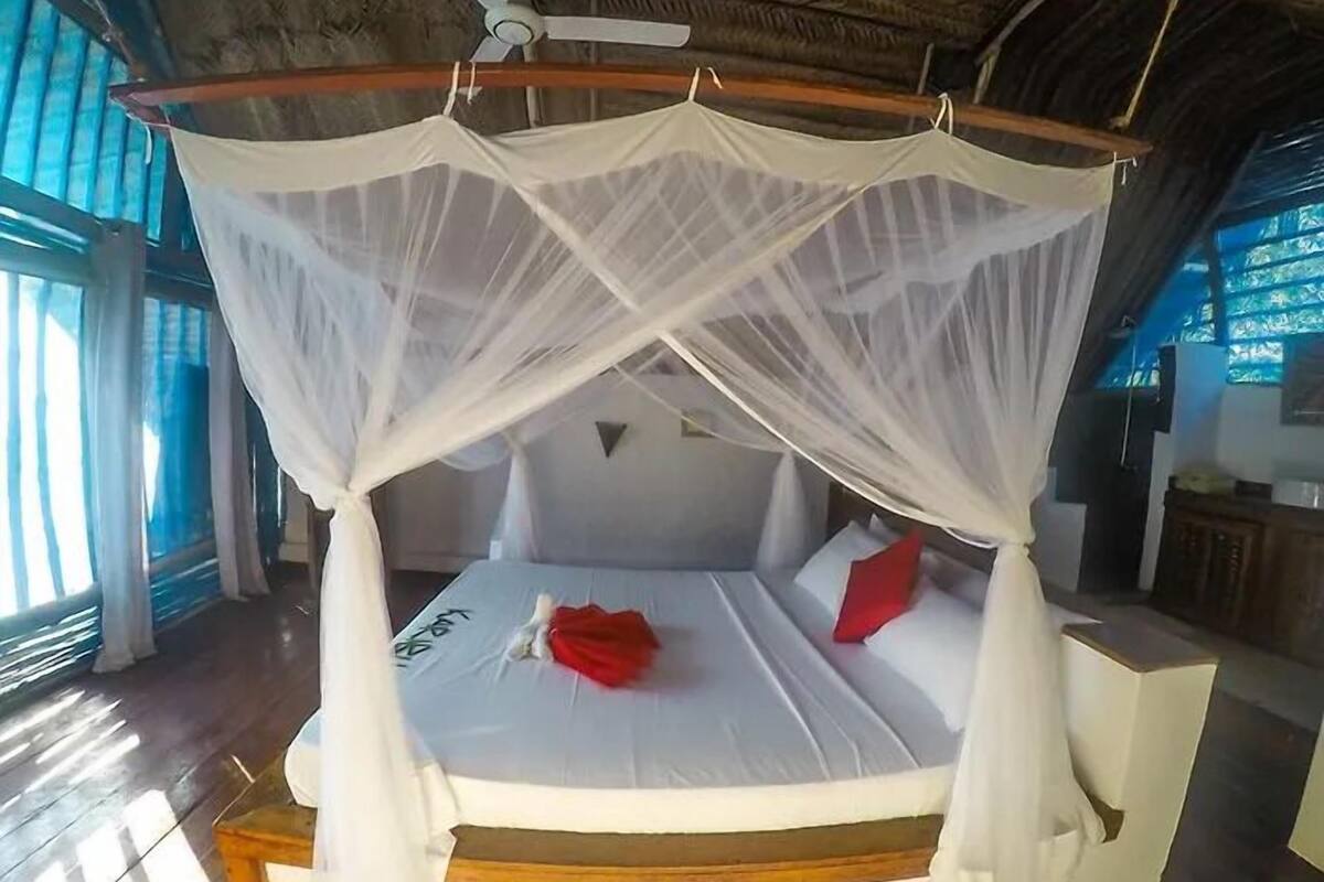 Una habitación con una cama bajo un dosel blanco, cabecera de madera y vistas al exterior a través de amplios ventanales.