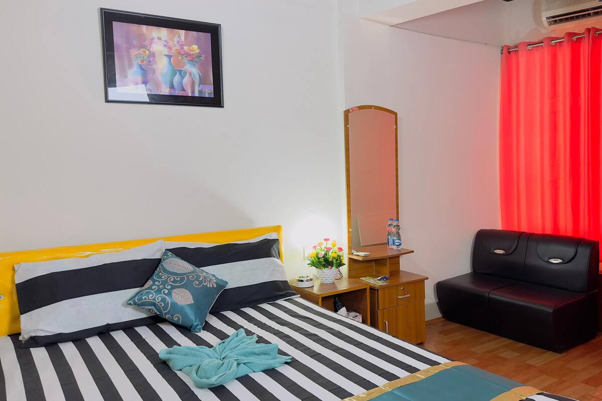 A hotel room with a bed, a desk with a mirror, a red curtain, and a black leather chair.
