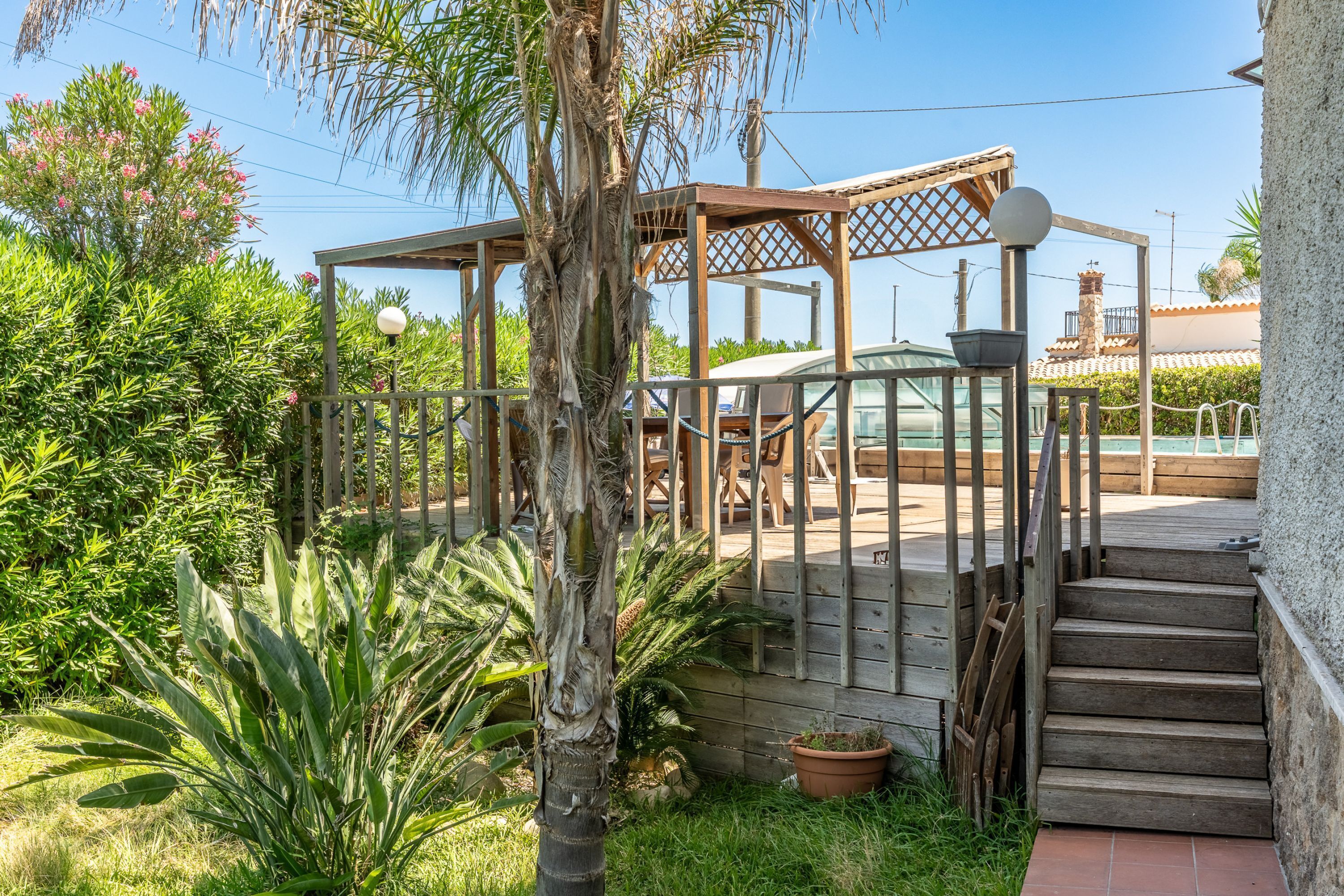 A wooden deck with a pergola, surrounded by lush greenery and a clear sky.