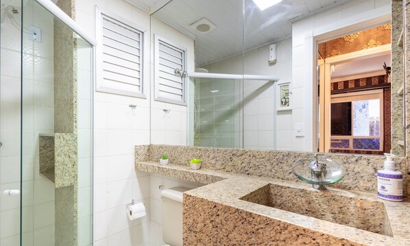 Un baño con encimera de granito, cabina de ducha de vidrio y un espejo que refleja una ventana con azulejos decorativos.