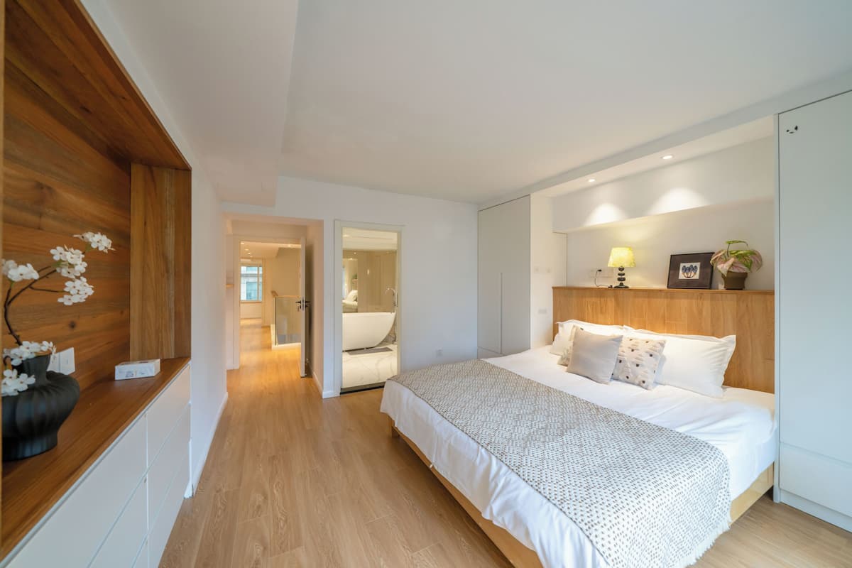 A bedroom with a wooden headboard, a bed with white bedding, a bedside table with a lamp, and a mirror above the bed.