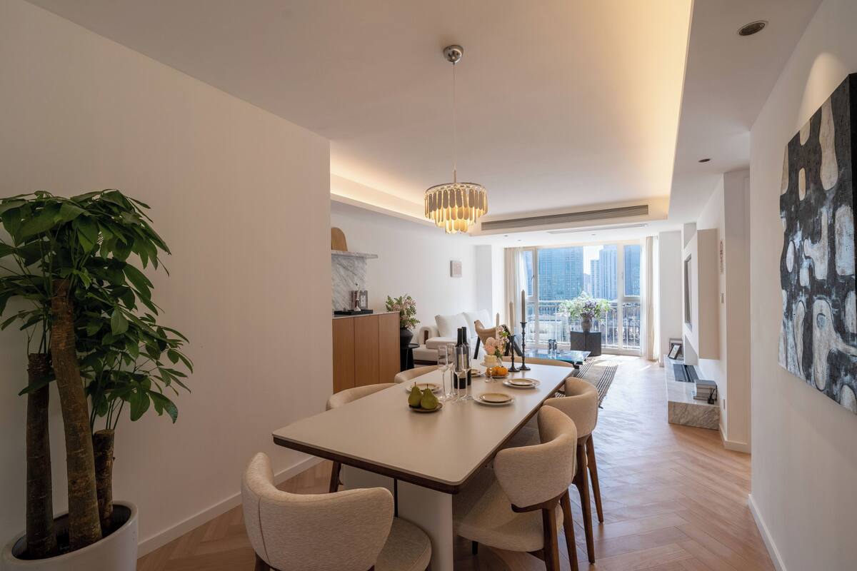 A modern dining area with a long table, chairs, a chandelier, and a large potted plant.
