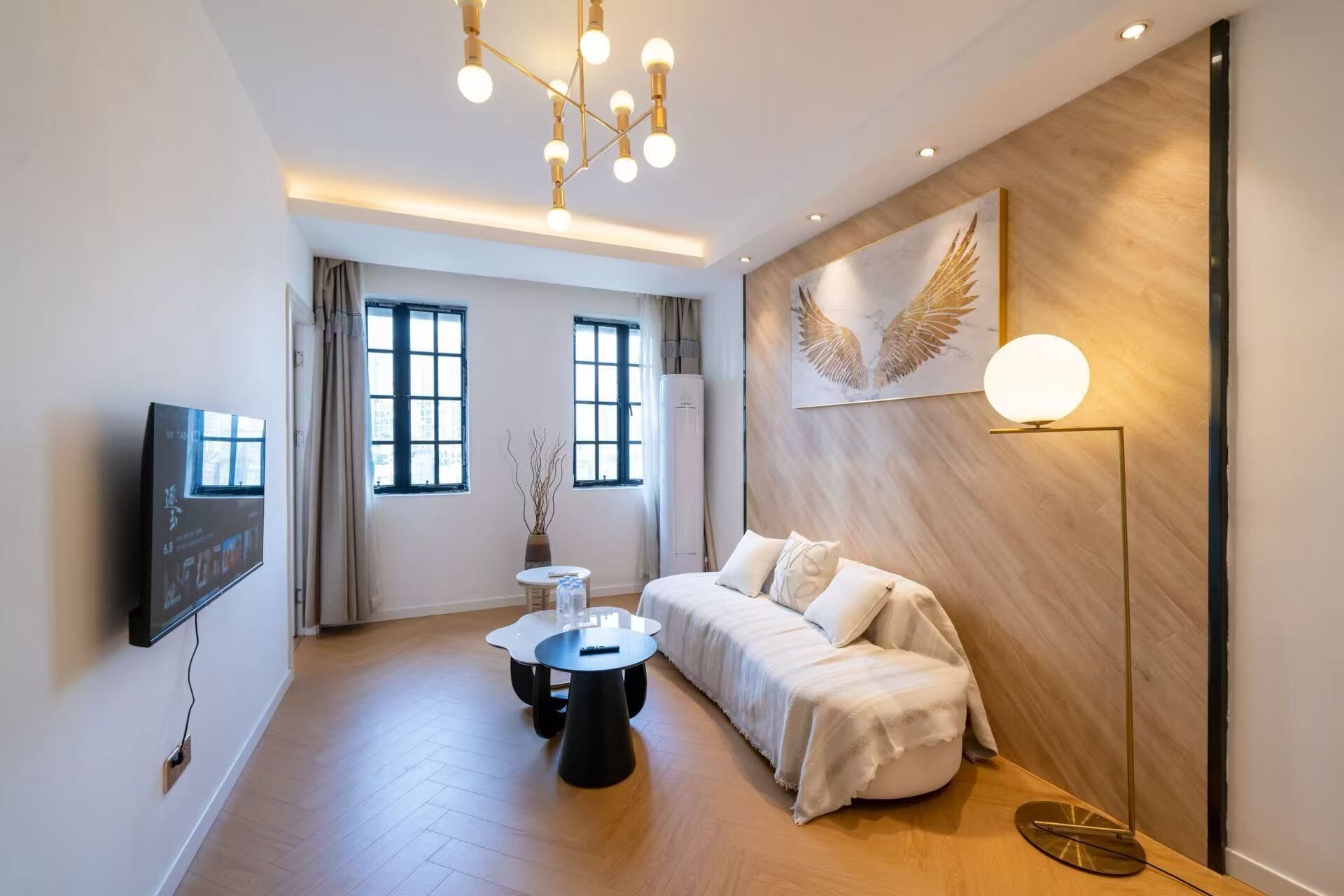A modern living room with a sofa, a coffee table, a TV mounted on the wall, and a large window.