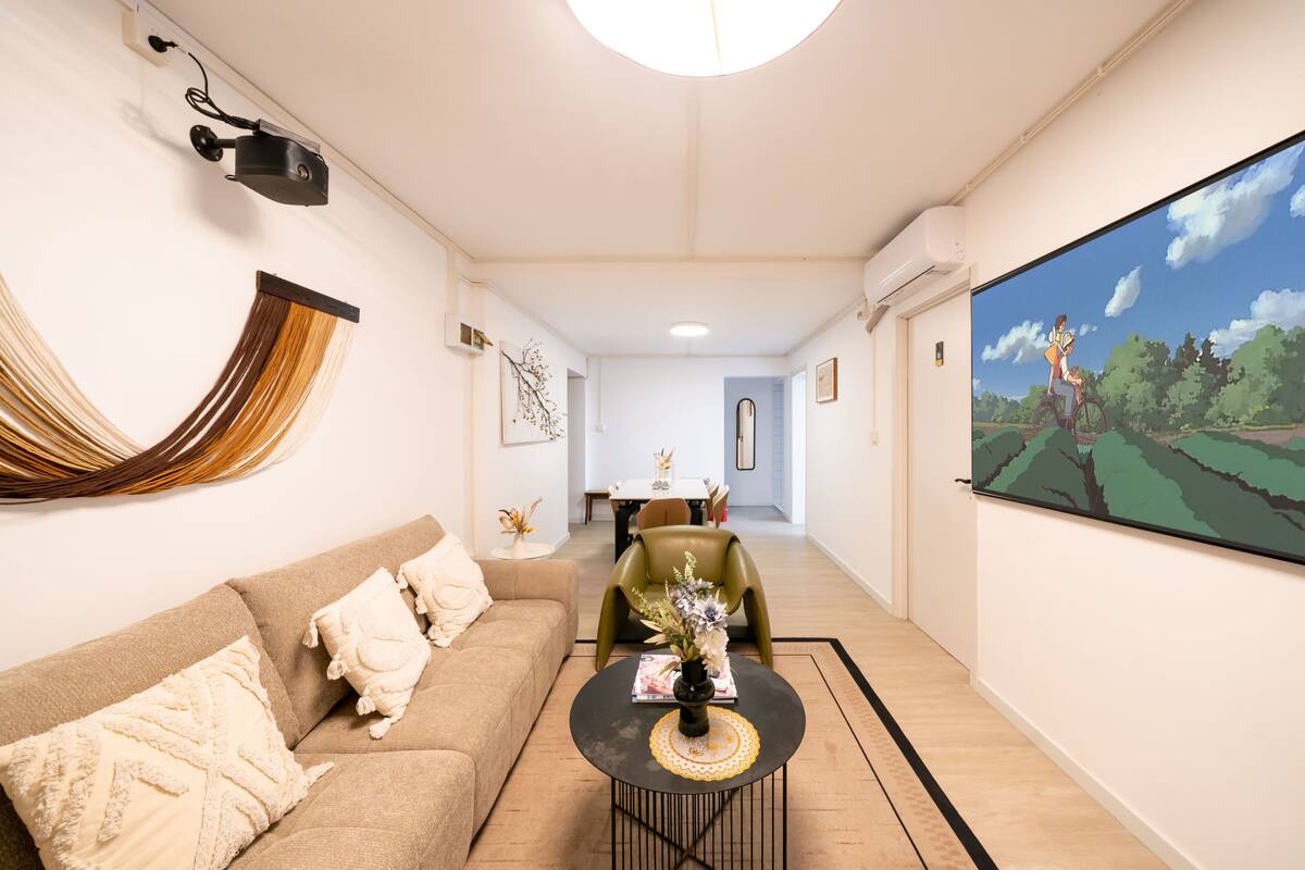 A modern living room with a sofa, a coffee table, a TV screen, and a dining area in the background.