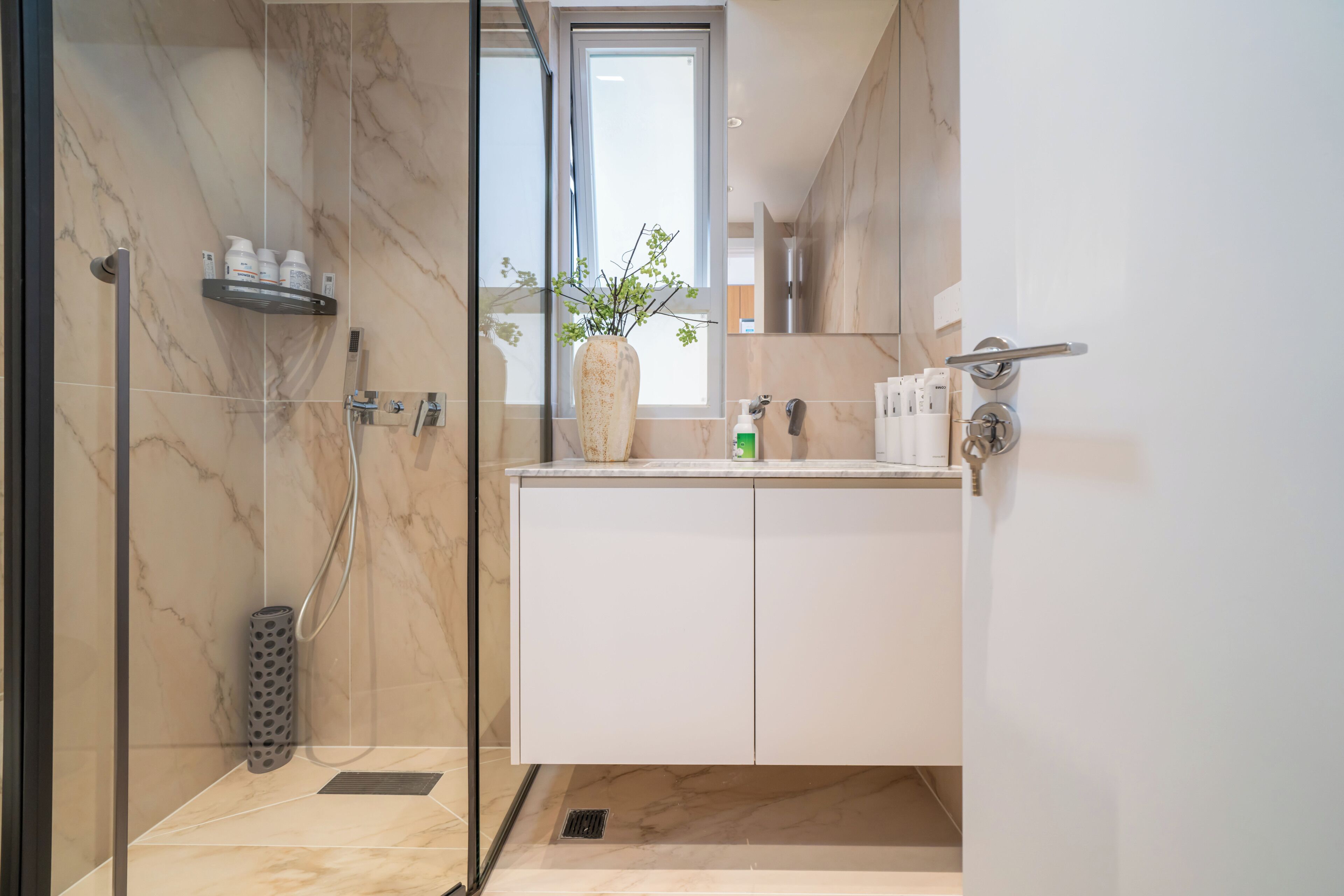 A modern bathroom with a glass shower enclosure, a marble-tiled shower area, a white vanity with a mirror, and a potted plant on the counter.
