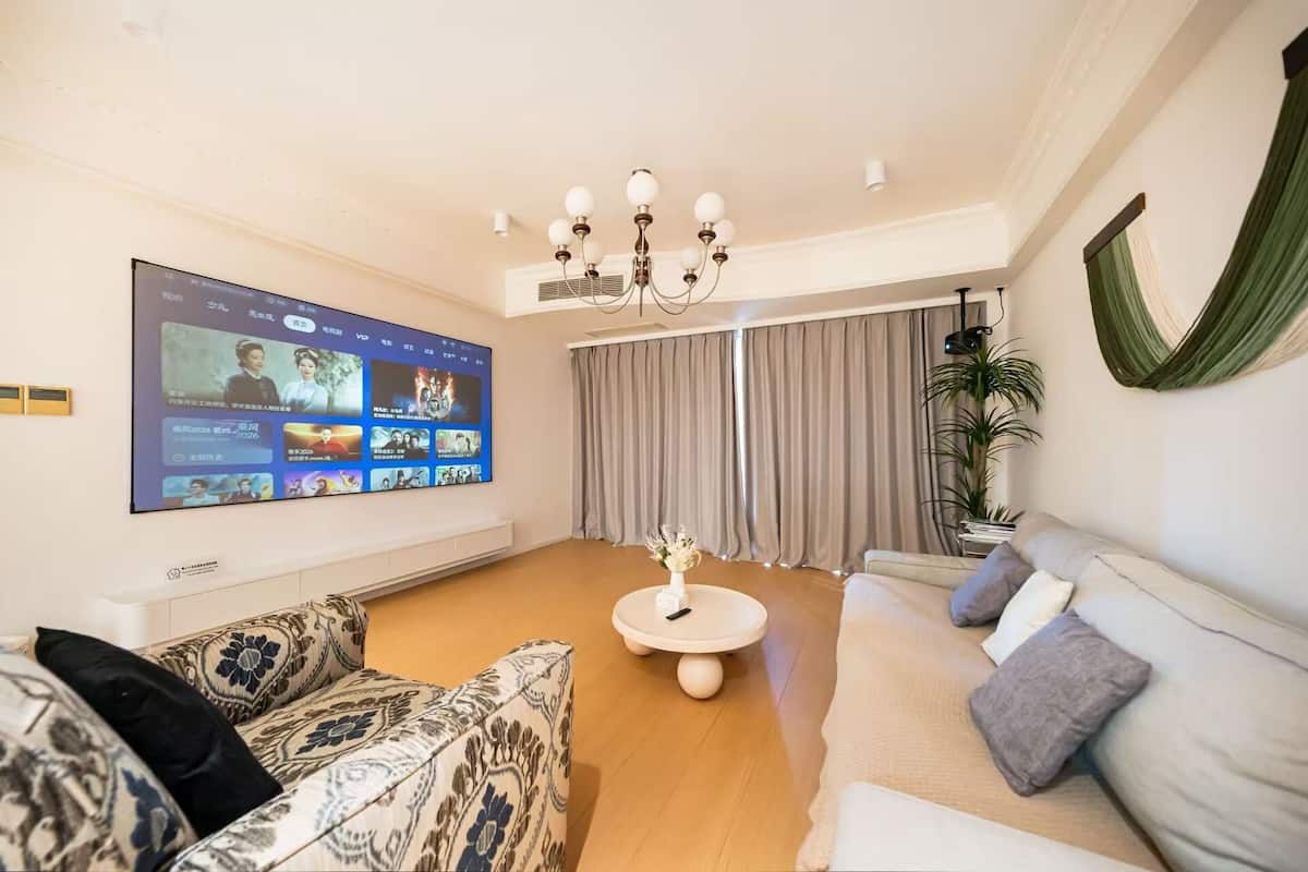 A living room with a large TV screen, a sofa, a coffee table, a chandelier, and a plant.