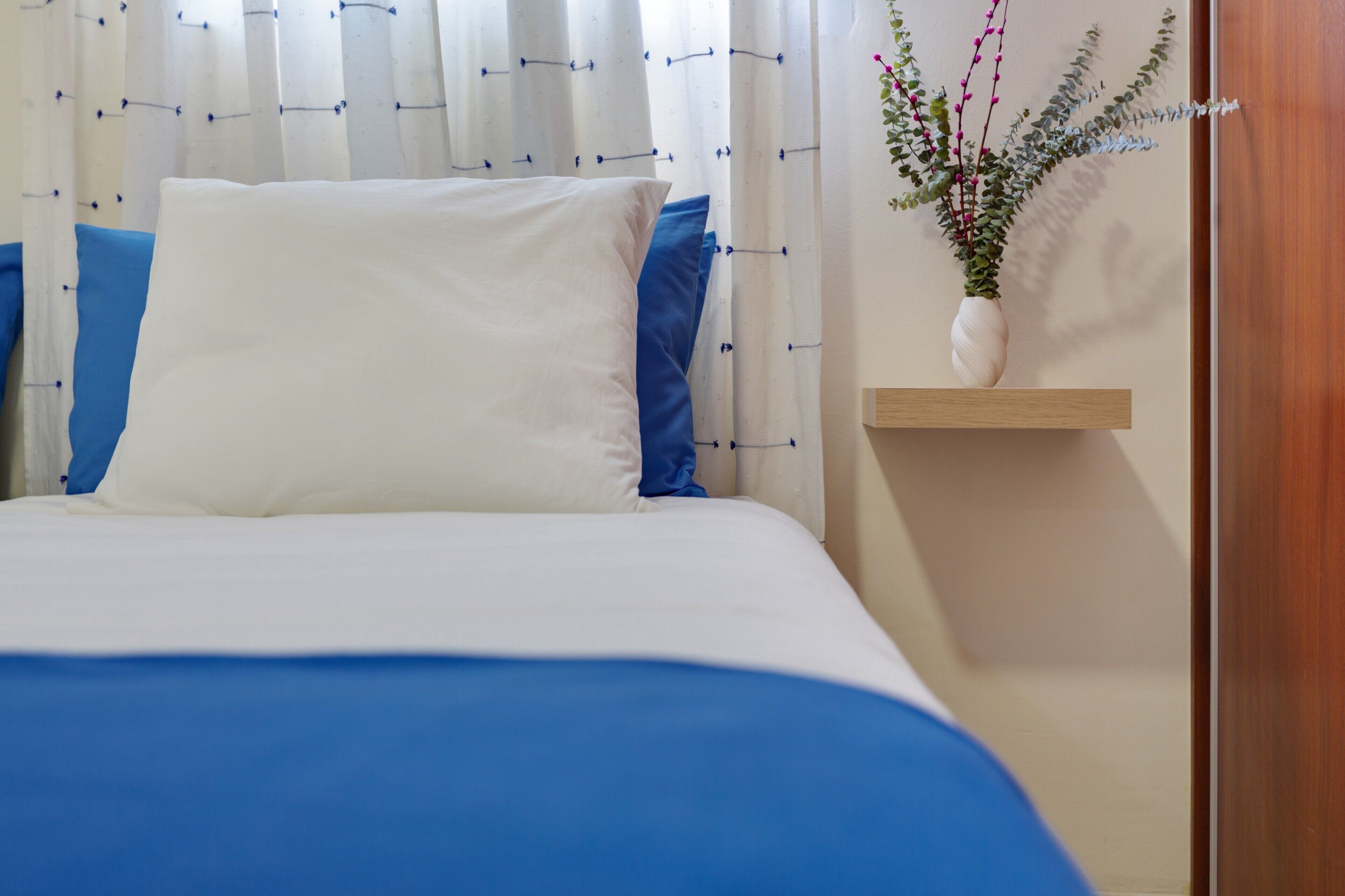 A bed with blue bedding, white pillows, and a white vase with flowers on a wooden shelf.