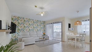 A living room with a sofa, coffee table, dining table, and chairs. There is a TV mounted on the wall, a ceiling fan, and a window with curtains. - Luxury Penthouse Plaza Apartment with Mountain View, Private Terrace, and Wi-Fi (Playa San Juan)