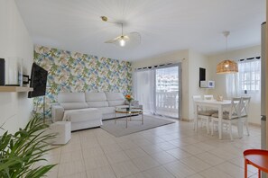 A living room with a sofa, coffee table, dining table, and chairs. There is a TV mounted on the wall, a ceiling fan, and a window with curtains.