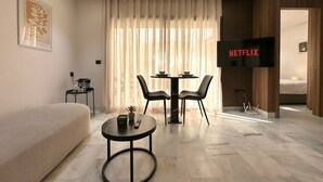 Una habitación de hotel moderna con una cama grande, un sofá, una mesa de comedor con sillas, un televisor que muestra 'NETFLIX' y una mesita de noche con una planta.