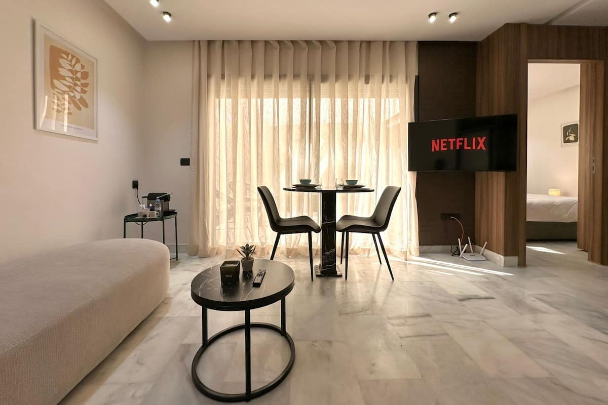A modern hotel room with a large bed, a sofa, a dining table with chairs, a TV displaying 'NETFLIX', and a small side table with a plant.