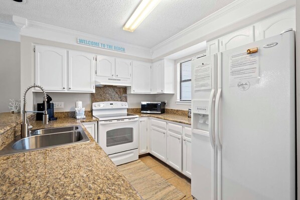 A kitchen with white cabinets, a granite countertop, a stainless steel sink, a stove, a microwave, and a refrigerator.