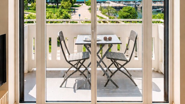 A balcony with a table and chairs, a TV, and a view of a lake and mountains. - Le Nouveau Rivage (T2 lake view) (Aix-les-Bains)