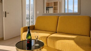 A modern living room with a yellow sofa, a glass table with a bottle and glass, and a view of the outdoors through large windows. - Le Nouveau Rivage (T2 lake view) (Aix-les-Bains)