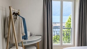 Ein Hotelzimmer mit einem Bett, einem Kleiderhaken aus Holz mit Kleidung, einem runden Tisch mit einem Stuhl und Meerblick durch ein mit Vorhängen drapiertes Fenster.