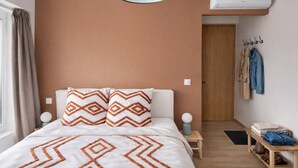 Ein Schlafzimmer mit Bett, Nachttisch, Lampe, Wandklimaanlage, Kleiderhaken mit Kleidung und einer Tür.