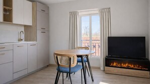 A modern kitchen with white cabinets, a wooden dining table, and chairs. A flat-screen TV and a fireplace are visible. - L'Ecume des Ondes (T2 mountain view) (Aix-les-Bains)