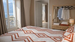 A hotel room with a bed, a wardrobe with hanging clothes, a bedside table with towels, and a view of the sea through large windows. - Les Flots Harmonieux (30m² lake view) (Aix-les-Bains)