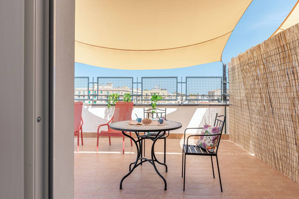 Un balcone con un tavolo rotondo, due sedie, un tendaggio giallo e vista sui palazzi.