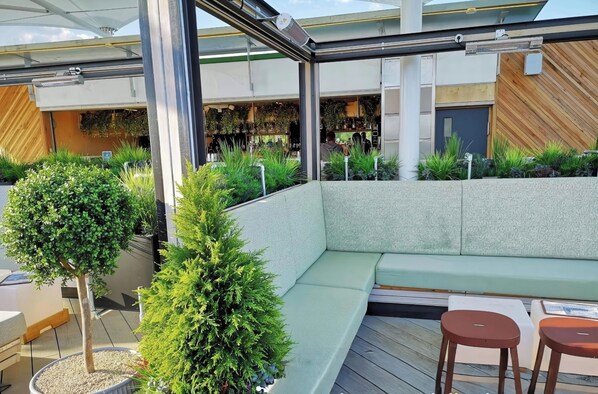 A modern outdoor seating area with a cushioned bench and two stools, surrounded by potted plants and a wooden deck.