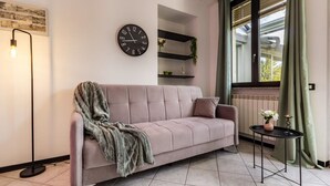 A modern living room with a sofa, a clock, a floor lamp, and a window with curtains. - Volta hub- parking lot and garden Lake Como 7min (Como)