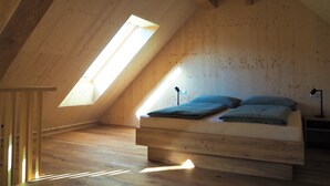 Une chambre à coucher aménagée dans un grenier, avec un plafond en pente, des poutres en bois, une lucarne, un lit avec des oreillers et une table de chevet.