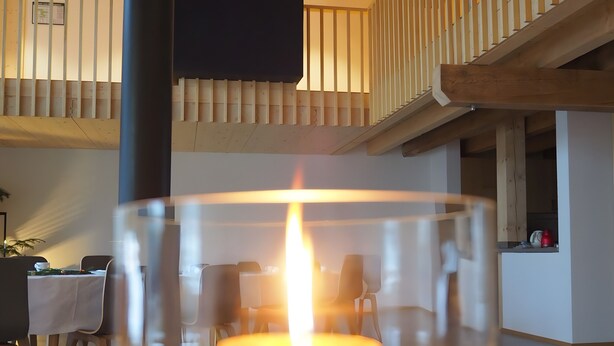A candle in a glass holder on a table with a wooden ceiling and a staircase in the background.