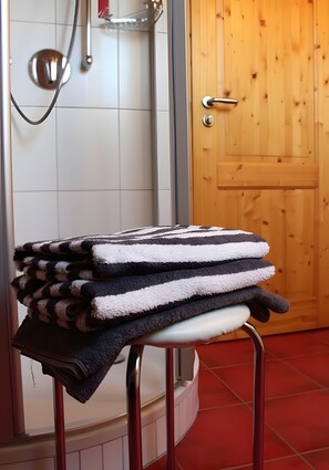 A bathroom with a shower, a wooden door, and a stack of towels on a stool.