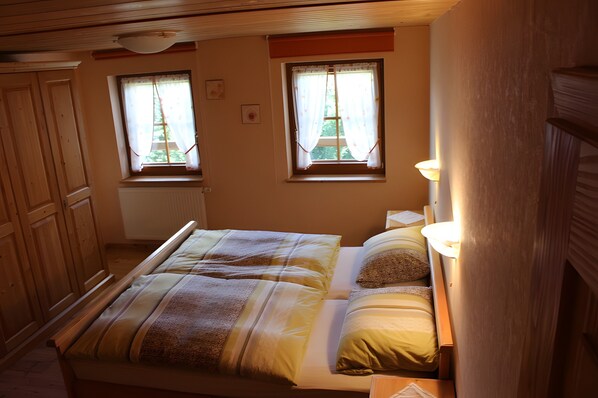 A bedroom with a bed, two windows with curtains, a radiator, wooden furniture, and a wall-mounted light fixture.