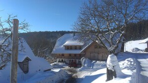A snow-covered rural house with a sloped roof, a wooden fence, and a clear blue sky.