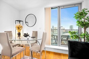 A modern dining area with a glass table, four chairs, a vase with dried flowers, a round mirror, and a view of the city through a large window.