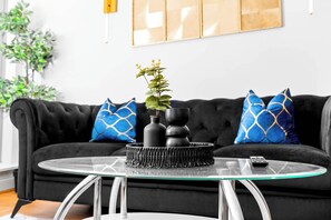 A modern living room with a black sofa, blue patterned cushions, a glass coffee table, and a potted plant.