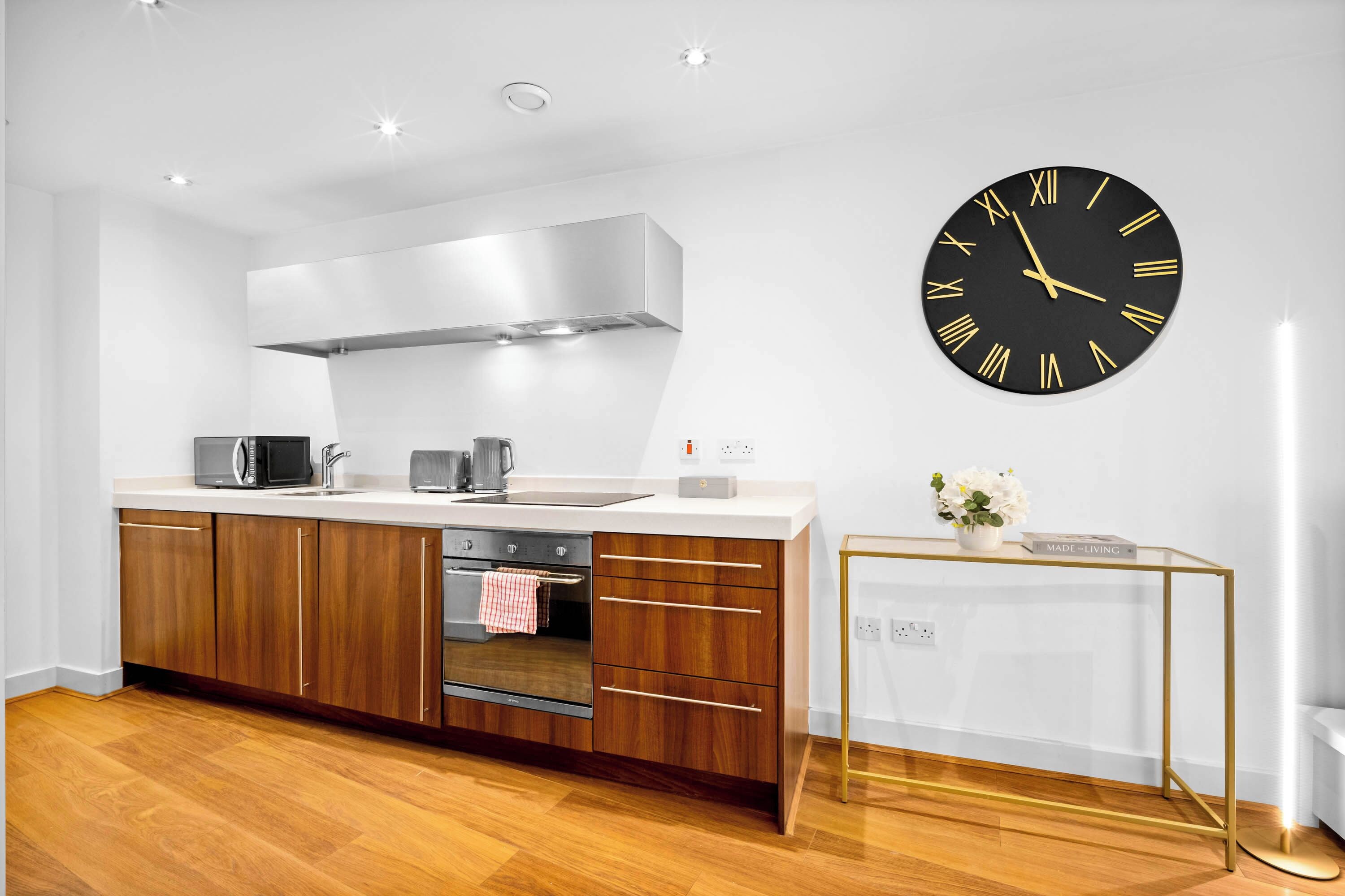 A modern kitchen with a large wall clock, wooden cabinets, and a small side table with a plant.