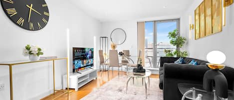 A modern living room with a large wall clock, a flat-screen TV, a sofa, a coffee table, and a view of the city through a large window.