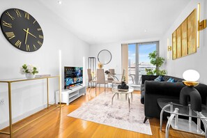 A modern living room with a large wall clock, a flat-screen TV, a sofa, a coffee table, and a view of the city through a large window.