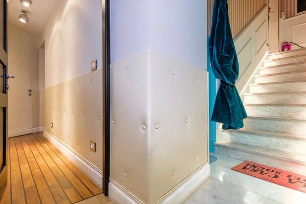 Un couloir doté d’un plancher en bois, d’un mur blanc et beige orné d’un motif en relief, et d’un escalier surmonté d’un rideau turquoise.